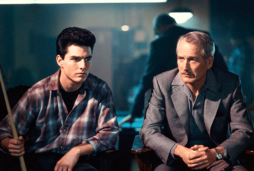 Tom Cruise, sentando e segurando um taco de sinuca, ao lado de Paul Newman no filme a cor do Dinheiro (1986). Em uma lista com os filmes de Tom Cruise.